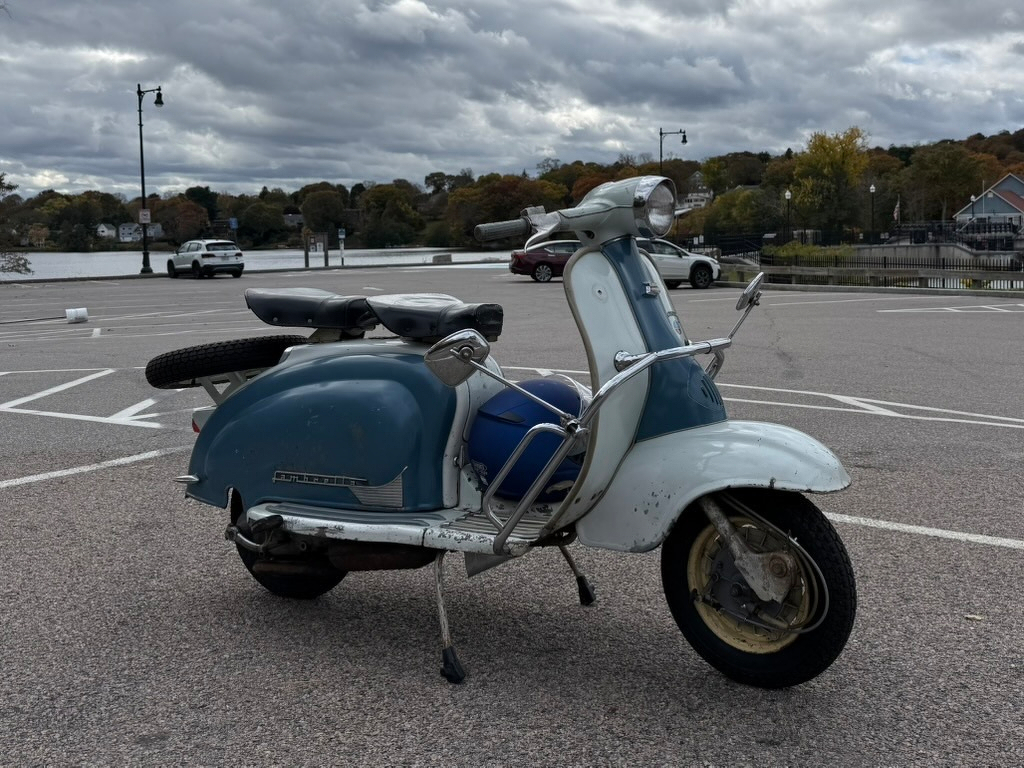 1962 blue and white Lambretta scooter in a parking lot, lake and treeline, with blue sky and clouds in the rear.
